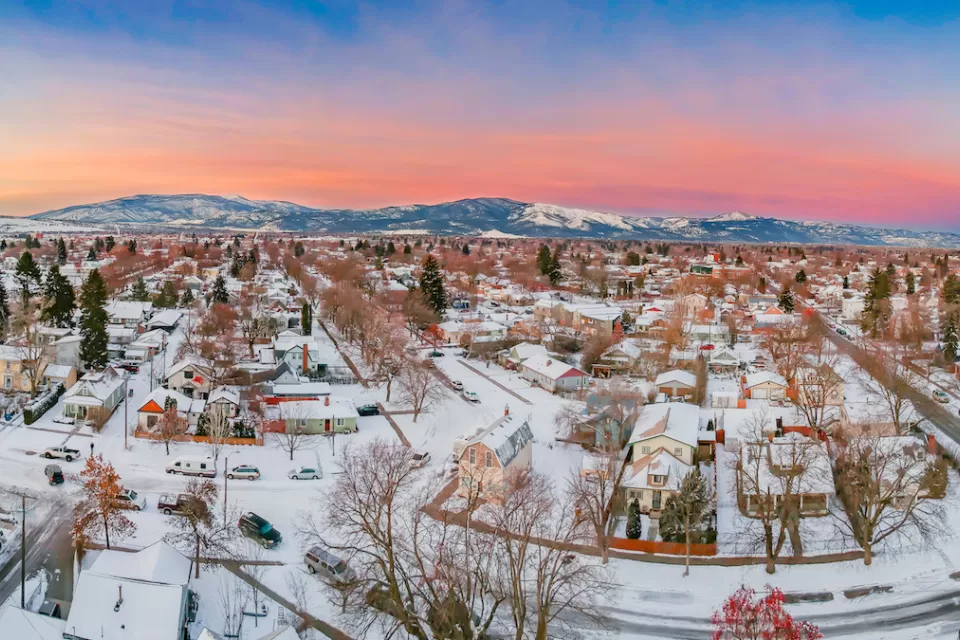 Large homes in neighborhood covered in snow in Missoula, Montana with mountains in background gainst sunset sky.